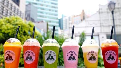 A colorful assortment of six Joe & The Juice smoothies in various flavors is displayed on a wooden surface outdoors. The flavors range from yellow to green to red to pink, each in a branded cup with a black straw. Urban buildings and greenery are in the background.