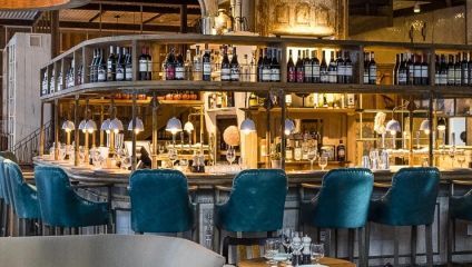 The image shows an upscale bar with teal upholstered barstools lined along a curved wooden bar counter. Above the counter, shelves display an array of wine bottles. Warm lighting lends a cozy ambiance to the interior decorated with wooden elements and metallic accents.