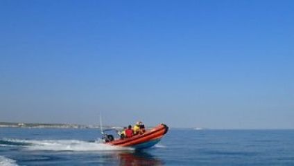 A small orange boat with people on board speeds across a calm blue sea, creating a white wake behind it. The sky is clear with no clouds visible. The horizon shows a faint outline of a coastline in the distance.
