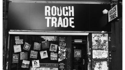 Black and white photo of the front of a Rough Trade record store. The facade has large text saying ROUGH TRADE. The window displays vinyl records, posters, and flyers. An additional hanging sign also reads ROUGH TRADE. The exterior appears worn and weathered.