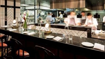 An upscale restaurant kitchen with chefs in motion, wearing white aprons, preparing meals. The foreground features a polished black counter with place settings, including white napkins, plates, and glassware. The decor includes black and white tiles and a flower vase.