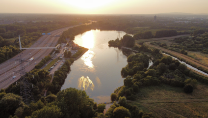 Aerial view of a river flanked by lush greenery and a road on the left during sunset. Several cars are parked along the road. The sun's reflection shimmers on the calm water, creating a peaceful ambiance. Power lines and distant buildings are visible in the background.