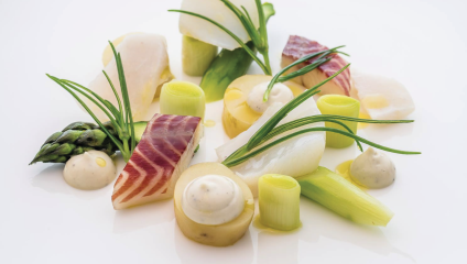 A gourmet dish of assorted fish fillets, thinly sliced vegetables, and small dollops of sauce elegantly arranged on a white plate. The presentation includes asparagus spears, leek slices, and garnishes of fresh green herbs.