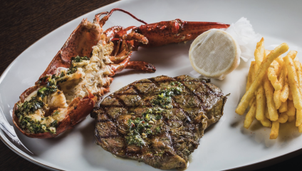 A neatly plated meal featuring a grilled lobster split in half, topped with herbs, next to a grilled steak garnished with a herb sauce. A small pile of French fries and a piece of lemon wrapped in cheesecloth accompany the dish, all served on a white plate.