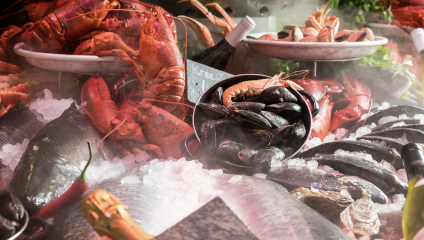 An assortment of fresh seafood displayed on a bed of ice. Lobsters, mussels, clams, oysters, and a variety of fish are arranged with some wine bottles nestled among them. The vibrant colors and freshness of the seafood are highlighted by the ice and surrounding greenery.