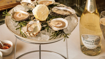 A platter of fresh oysters served on a bed of ice is garnished with a lemon slice and green herbs. Beside the oysters, there's a bottle of Perrier-Jouët Blanc de Blancs champagne and a glass of champagne. Small bowls of condiments are visible in the background.