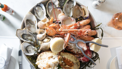 A seafood platter filled with oysters, shrimp, crab, and other shellfish, garnished with seaweed and lemon halves. Silverware and napkins are placed around the platter on a white table. Bottles of hot sauce and salt and pepper shakers are nearby.