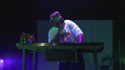 A person in a white shirt and bandana stands behind a table on a dimly lit stage, thoughtfully touching their chin. The table has some plants and small jars on it. The background is dark with a subtle purple and green hue.