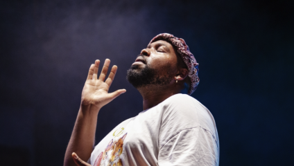 A person wearing a white T-shirt and a head wrap stands with eyes closed and hand raised, immersed in a moment under dramatic lighting. The background is dark and slightly smoky, creating a focused and contemplative atmosphere.