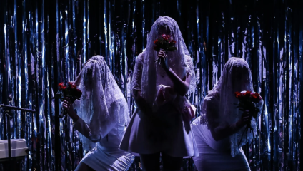 Three individuals in white outfits and veiled headpieces hold red bouquets, standing on a dimly lit stage. The backdrop is adorned with metallic, shimmering curtains. The mood is theatrical and mysterious.