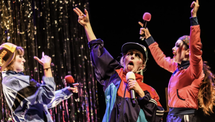 Three people dressed in vibrant 90s-inspired clothing enthusiastically perform on stage under shiny, metallic streamers. The central figure wears a hat and appears to sing passionately into a microphone while making a peace sign. The other two also hold microphones, singing or cheering.