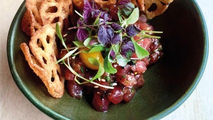 A green bowl contains a dish of tuna tartare topped with a quail egg yolk and microgreens. To the side, there are crispy, lattice-patterned lotus root chips. The presentation is elegant, with vibrant colors from the fresh ingredients.