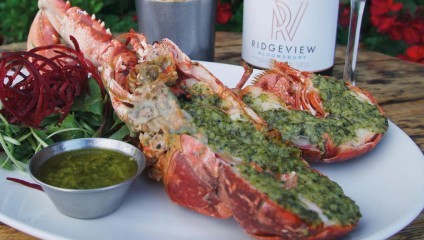 A plate with a grilled lobster topped with a green herb sauce, accompanied by a small dish of extra sauce, a fresh salad with beet spirals, and a bottle of Ridgeview Bloomsbury. The dishes are set on a wooden table with a backdrop of red flowers.