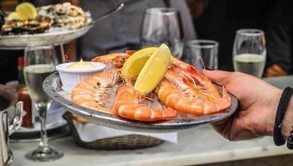 A person holds a metal plate with large cooked shrimp, lemon wedges, and a small dipping sauce container. In the background, there are glasses of white wine and a basket of bread, with more food and diners visible.