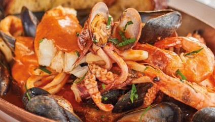 A close-up of a seafood dish featuring mussels, shrimp, squid, and chunks of fish drenched in a rich red sauce, garnished with chopped herbs. The dish is served in a copper pan, with a piece of bread visible in the background.