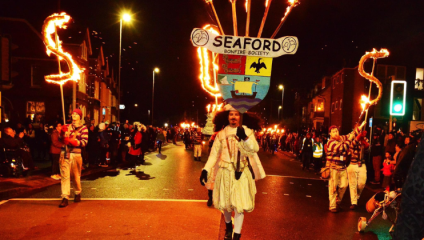 Torchbearers from the Seaford Bonfire Society walks in procession through a street in Seaford while spectators watch from the pavement on either side. The leading torchbearer is dressed in a traditional white period costume, and carries the banner of the Seaford Bonfire Society. Behind him on both sides, torchbearers carry a flaming S, and are each wearing a red beanie, a red-and-white striped jumper and white trousers.