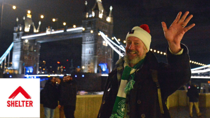 A bearded man wearing a red Santa hat waves at the camera, smiling as he walks the Walk for Home for homelessness charity Shelter. In the background, we see Tower Bridge against the night sky.