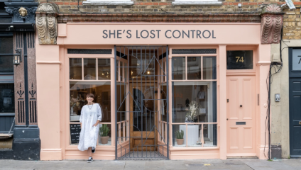 A woman in a white dress stands casually outside a boutique store named SHE'S LOST CONTROL. The storefront is painted in pastel pink with large windows displaying various items. The door is partially open, and the building is located at number 74.