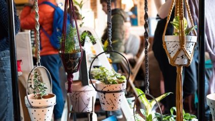 A lively indoor scene at a market or fair showcasing hanging potted plants. Each pot, holding various green plants, is suspended by different colored macramé hangers. Blurred people are seen in the background, creating a bustling atmosphere.