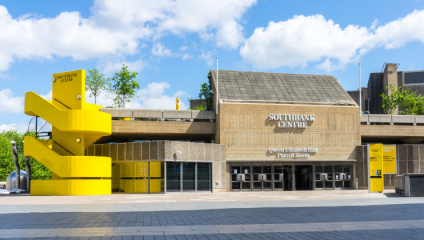 The Southbank Centre in London on a sunny day with a clear blue sky. The building showcases modern architecture with a prominent yellow spiral staircase on the left. The entrance signs indicate Queen Elizabeth Hall and Purcell Room.