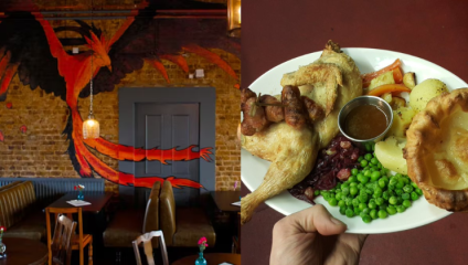 A hand holding a plate with roasted chicken, potatoes, peas, carrots, and a Yorkshire pudding in the foreground, with a small dish of gravy on the side. In the background, there's a restaurant with a painted mural of a phoenix on a brick wall.
