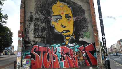 A large mural on the side of a building depicts a stylized face in yellow and black tones with red accents. The lower portion of the mural features bold, colorful graffiti text that reads STOKE against a gritty urban backdrop.