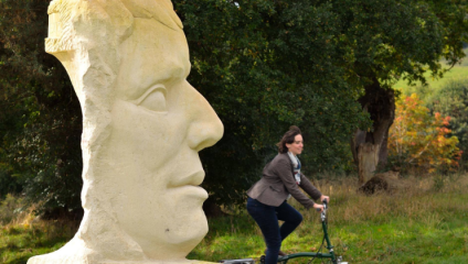 A woman rides a bicycle past a large stone sculpture of a human head in an outdoor setting. She is wearing a brown jacket and a scarf. The background features greenery and trees.
