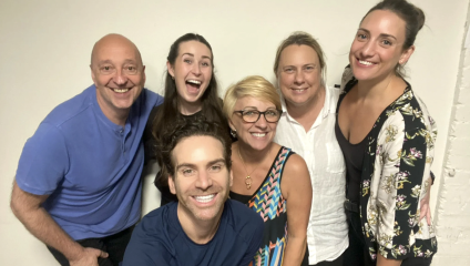 A group of six people, three in the front and three in the back, are smiling and posing for a photo against a plain wall. They appear to be happy and enjoying each other’s company.