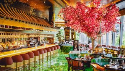 A luxurious restaurant interior featuring a large, vibrant cherry blossom tree sculpture near the center. The bar area on the left has plush pink stools, golden fixtures, and mirrored walls, while the seating area on the right has elegant green and pink chairs with lavish decor.