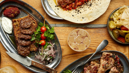 A top view of a wooden table set with assorted dishes: a plate with sausage, sliced bread, tomatoes, herbs, and a dollop of sauce; a tortilla with minced filling; a bowl of pickled vegetables; a beverage with a frothy top; and a dish with cooked meat and green beans.