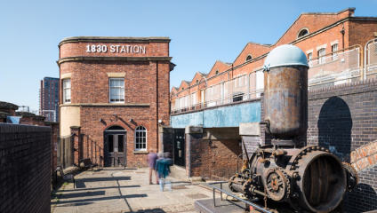 A scene of the historic 1830 Station featuring red brick buildings and an industrial setting. The station's sign is prominently displayed on a two-story building. There's an old, rusted steam engine in the foreground, with a few blurred figures walking nearby.