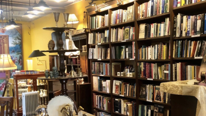 A cozy, well-lit bookstore with tall, wooden shelves filled with books. There are various lamps, decorative items, and a drum placed among the books and furniture. The walls are painted a warm yellow, contributing to the inviting atmosphere.