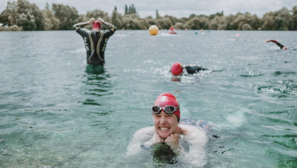 A person in a red swim cap and goggles smiles while resting on the water's surface in a lake. Behind, one swimmer adjusts their goggles while standing, and another swims. Trees and buoy markers are visible in the background under a cloudy sky.