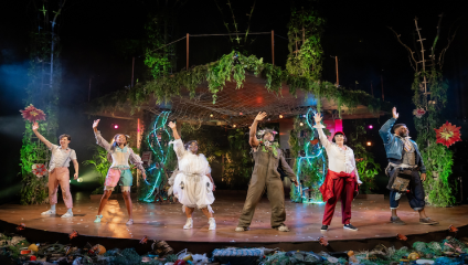 A diverse group of six actors stands on an outdoor stage, dressed in eclectic costumes and posing mid-bow with one arm raised each. The stage is decorated with lush greenery, flowers, and warm lighting, creating an enchanting forest-like atmosphere.