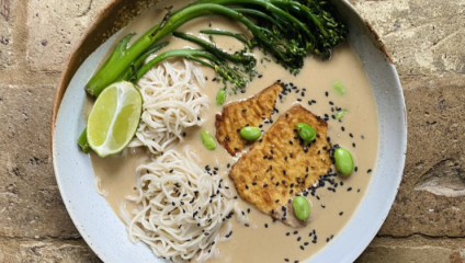 A bowl of creamy soup with noodles, tofu, broccolini, edamame, a lime wedge, and black sesame seeds on a rustic table. The tofu is pan-fried and placed prominently in the center. The dish has a light tan broth and is garnished with green vegetables.