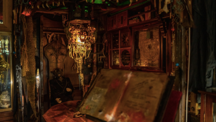 A dimly lit room filled with various antique and mysterious objects. A large, open, weathered book with indistinct text is situated in the foreground. In the background, there are ornate decorations, a dim light fixture, and an eerie figure dressed in dark clothing.