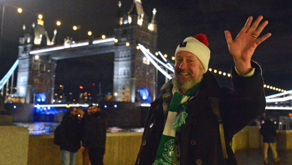 A bearded man in a Santa hat, taking part in the Walk for Home for the charity Shelter, waves at the camera with Tower Bridge in the background against the black night sky.