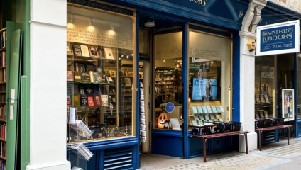 A quaint bookstore with a blue storefront displays an assortment of books. The store's windows exhibit various titles, while a sign above reads Watkins Books. A bench with more books is positioned outside, and a smaller hanging sign shows the store's contact number.