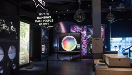 An exhibition room with a central display featuring a colorful circular chart. Above, a sign asks, Why do rainbows make people happy? Various interactive exhibits and information boards are arranged around the room, with dim lighting and decorative hanging lights.