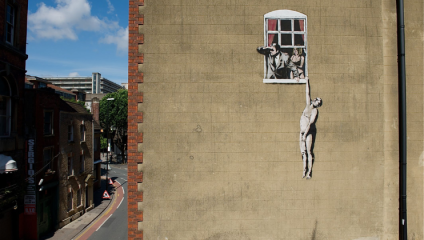 Mural on the side of a building depicting a naked man hanging out of a window, while a shocked woman looks on from inside and a man peers out from behind curtains. The artwork is on a brick wall with other buildings and a street visible nearby.