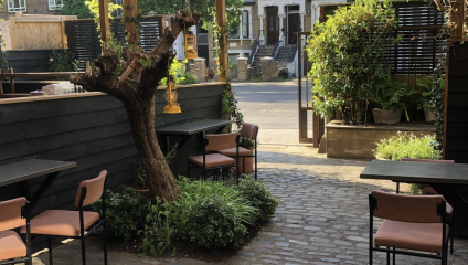 Outdoor cafe seating area with several tables and chairs, surrounded by greenery. A large tree trunk stands at the center, and wooden panels border the area. In the background, there is a quiet residential street with parked cars.