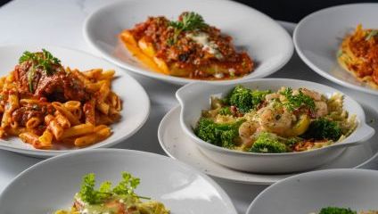 A selection of Italian dishes is displayed on a table, including pasta varieties like penne with tomato sauce and meatballs, lasagna, and pasta with shrimp and broccoli. Each dish is garnished with fresh herbs on white plates.