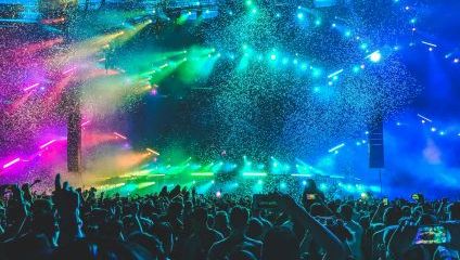 A vibrant concert scene with colorful stage lights in shades of green, blue, purple, and yellow illuminating the audience. A large crowd of people are cheering and raising their hands, capturing the moment with phones. Confetti is falling from above, enhancing the festive atmosphere.