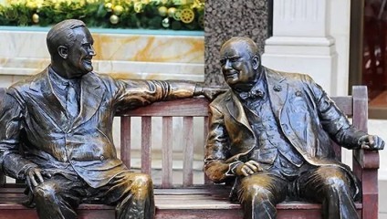 Bronze sculptures of two men, depicted sitting on a bench in conversation. The man on the left has his arm draped over the back of the bench while the man on the right sits with a relaxed posture. A decorated holiday display is visible in the background.