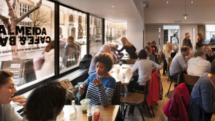 A bustling café with patrons engaged in conversations and enjoying their drinks. Large windows reveal a street with trees and buildings outside. People of various ages are seated at tables, some waiting in line, and the interior has a cozy, modern ambiance.