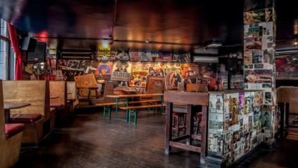 A dimly-lit room with wooden booths and tables, featuring numerous vibrant posters and photos covering the walls and pillars. The decor has a vintage, eclectic vibe, with images of musicians, bands, and retro advertisements. The floor is dark wood.