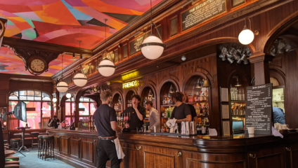 A lively bar with a colorful, geometric patterned ceiling. Three bartenders are engaging with customers behind a wooden counter adorned with hanging globe lights. The shelves behind the bar are stocked with various bottles and a neon sign that reads FRIENDS.