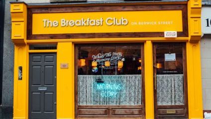 A vibrant storefront with yellow and wood accents displays The Breakfast Club on Berwick Street sign above. A neon sign reading Morning Glory is visible in the window, which has lace curtains. The entrance features a gray door to the left and wooden-framed windows.