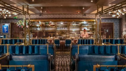 A sophisticated restaurant interior featuring plush blue booths, a mosaic-patterned carpet, and an ornate, well-stocked bar with golden accents. A blurred figure of a bartender is seen behind the bar, affecting an ambiance of luxury and elegance.