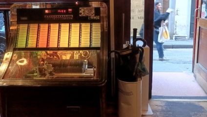 A vintage jukebox with colorful song selection panels is positioned near a doorway. Next to it is a beige umbrella stand holding several umbrellas. A person carrying a shopping bag walks past the open door on the sidewalk outside.